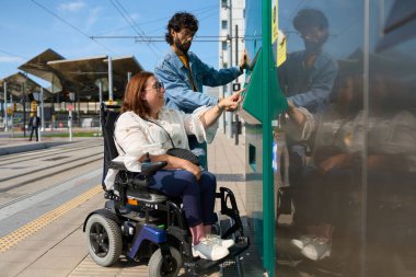 Engelli bir kadın, bir arkadaşının yardımıyla tramvay veya tren istasyonunda bilet otomatı kullanıyor, erişilebilirliği ve dahil edilmeyi teşvik ediyor.