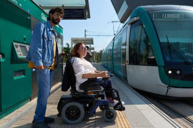 Tekerlekli sandalyedeki kadın istasyonda yol arkadaşıyla tramvay yardımı bekliyor.