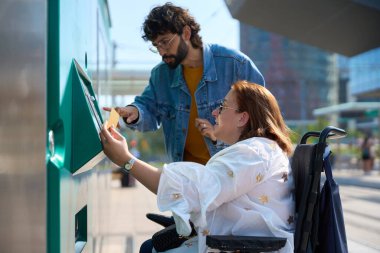 Engelli bir kadın tren istasyonunda arkadaşına yardım ederken bilet otomatı kullanıyor.