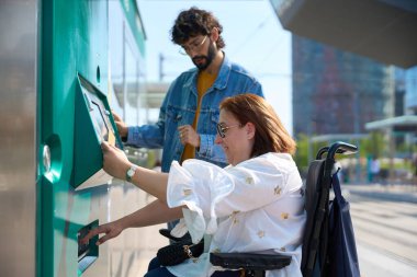 Engelli bir kadın toplu taşıma istasyonunda bilet otomatı kullanıyor, bir arkadaşın yardımıyla, erişilebilirliği ve dahil edilmeyi teşvik ediyor.