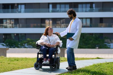 Doktor engelli bir kadına parkta tekerlekli sandalyede yardım ediyor, onunla konuşuyor ve tıbbi destek sağlıyor.