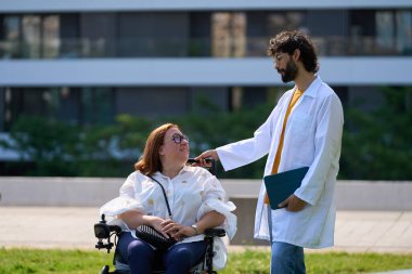 Tekerlekli sandalyede oturan engelli bir kadını destekleyen doktor park ortamında yardım ve empati teklif ediyor.