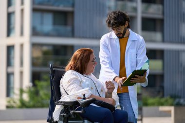 Doktor, parkta tekerlekli sandalyede oturan gülümseyen bir hastaya tıbbi belgeler gösteriyor.