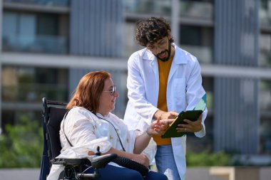 Doktor, güneşli bir günde tekerlekli sandalyedeki gülümseyen bir kadına tıbbi bilgileri açıklıyor.