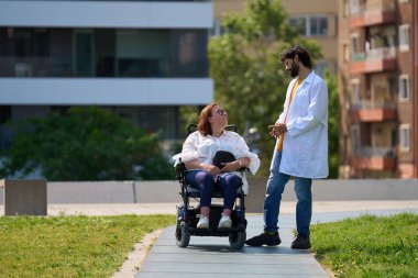 Doktor, dışarıda parkta elektrikli sandalyede oturan bir kadın hastayla konuşuyor.