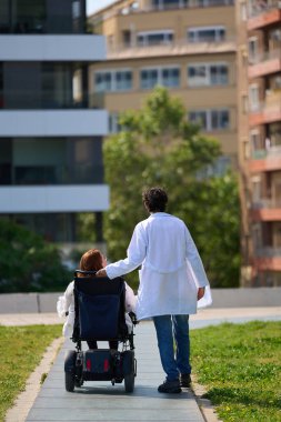 Doktor tekerlekli sandalyedeki kadın hastaya yardım ediyor, şehir parkında kaldırımlı bir yolda yol alıyor, bakım ve hareketliliği sembolize ediyor.