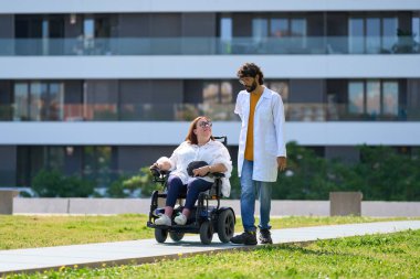 Elektrikli sandalyeli engelli bir kadına yardım eden doktor güneşli bir parkta yürüyüşün tadını çıkarıyor, kapsayıcılığı ve ulaşılabilirliği teşvik ediyor.