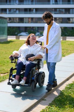 Doktor elektrikli sandalyeli neşeli engelli bir kadına yardım ediyor, güneşli bir günün tadını çıkarıyor.