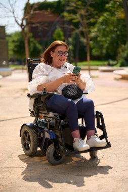 Parkta elektrikli sandalyede otururken cep telefonu kullanan engelli mutlu bir kadın.