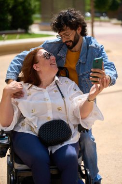 Gülümseyen engelli kadın erkek arkadaşıyla selfie çekiyor, güneşli bir günde parkta eğleniyor.