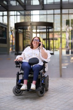 Tekerlekli sandalye kullanan ve şehir ortamında akıllı telefondan konuşan mutlu bir iş kadını.