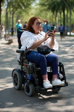 Parkta tekerlekli sandalyeyle gezerken akıllı telefon kullanan mutlu bir kadın.