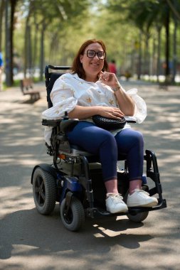 Elektrikli sandalyesini şehir parkında kullanan engelli mutlu kadın güneşli bir günün tadını çıkarıyor.