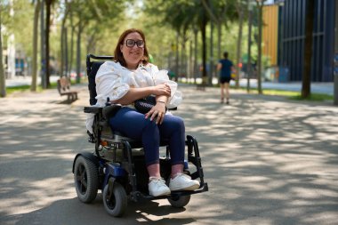 Elektrikli sandalye kullanan mutlu kadın parkta güneşli bir günün tadını çıkarıyor.