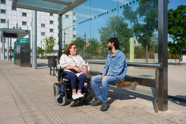 Tekerlekli sandalye kullanan bir kadın otobüs durağında bir adamla konuşuyor, arkadaşça bir sohbetten hoşlanıyor.