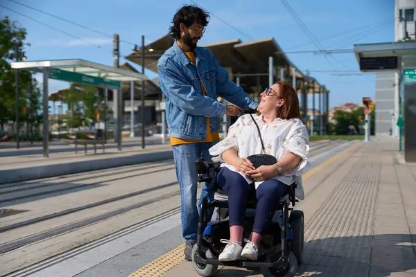 Değer veren bir adam, modern bir tramvay istasyonunda bekleyen gülümseyen engelli bir kadının omzunu düzeltir.