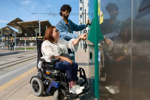 Engelli bir kadın, bir arkadaşının yardımıyla tramvay veya tren istasyonunda bilet otomatı kullanıyor, erişilebilirliği ve dahil edilmeyi teşvik ediyor.
