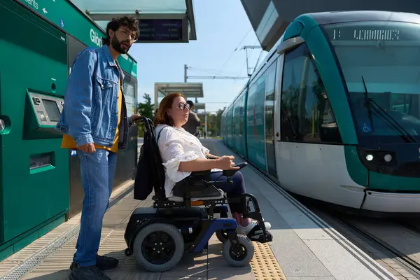 Tekerlekli sandalyedeki kadın istasyonda yol arkadaşıyla tramvay yardımı bekliyor.