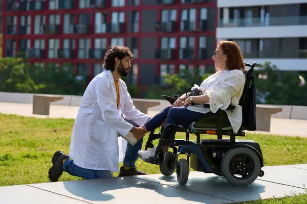 Parkta tekerlekli sandalyesinde oturan bir kadın hastanın bacağını muayene eden doktor engelli insanlara ilgi ve destek gösteriyor.
