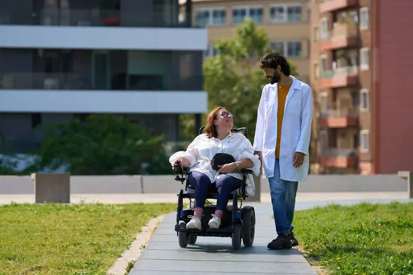 Elektrikli sandalyede sakat bir kadına yardım eden doktor güneşli bir şehir parkında birlikte yürümekten zevk alıyor.