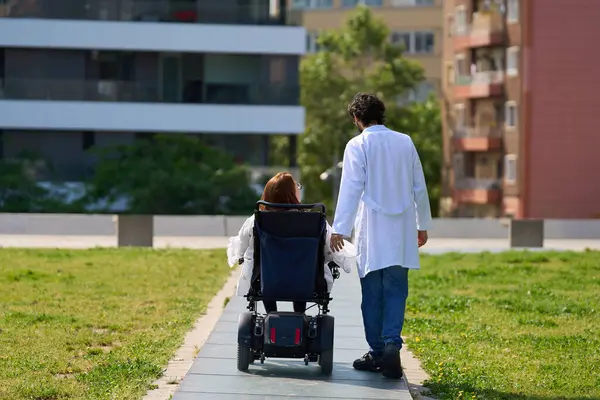 Elektrikli sandalyede sakat bir hastaya yardım eden doktor güneşli bir şehir parkında geziniyor.