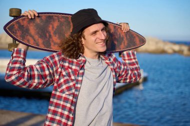 Kıvırcık saçlı, gülümseyen ve kıyı boyunca yürüyen, omuzlarında uzun bir tahta taşıyan, güneşli sahil kıyısının keyfini çıkaran genç bir adam.