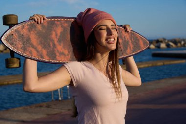 Genç bir kadın gülümsüyor ve omuzlarında bir uzun tahta tutuyor, güneşli deniz ve canlı kumsalda kaygısız anların tadını çıkarıyor.