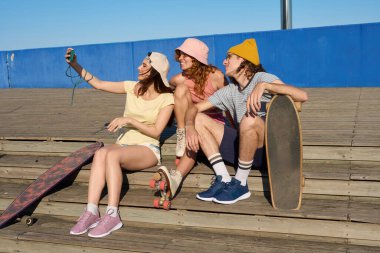 Üç genç kaykaycı arkadaş güneşli bir günde ahşap merdivenlerde otururken akıllı telefonlarıyla selfie çekiyorlar.