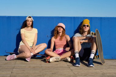 Üç havalı genç patenci ahşap bir geçitte canlı mavi bir duvarın karşısında güneşli havanın tadını çıkarıyorlar.