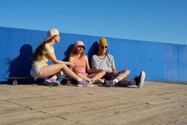 Üç patenci ahşap bir geçitte oturmuş, canlı mavi bir duvara karşı birbirlerine eşlik ederek eğleniyorlar.