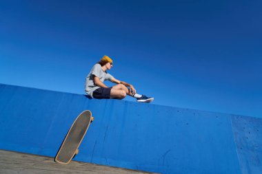 Genç patenci parlak, berrak bir gökyüzünün altında parlak mavi bir duvara yaslanmış, kaykayı yakınlarda rastgele eğilmiş.