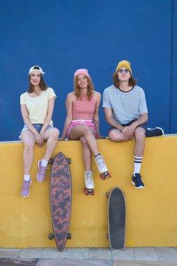 Üç havalı genç sarı bir duvarda mavi arka planda oturuyorlar, kaykay ve patenleriyle poz veriyorlar.