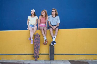 Üç genç arkadaş kaykayları ve patenleriyle takılıyor, canlı mavi ve sarı bir duvara karşı mola veriyorlar.