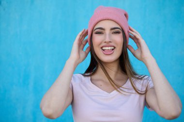 Neşeli, şık bir kadın pembe beresini düzeltirken parlak mavi bir arka plana gülümsüyor, gençlik mutluluğu yayıyor.