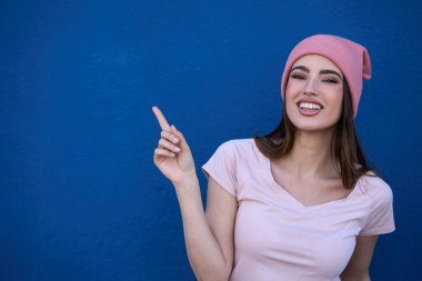 Gülümseyen kadın, parlak mavi bir arkaplanı işaret ederek neşeli ve pozitif bir ifade sergiliyor.