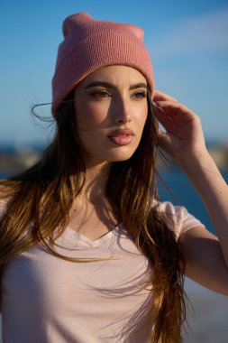 Pembe bereli şık genç bir kadın uzun kahverengi saçlarını güneşli ve rüzgarlı bir plajda düzeltir.