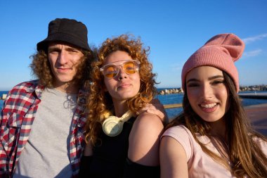 Bir grup neşeli genç arkadaş mükemmel bir yaz gününde güneşli bir sahilde neşeyle kucaklaşıp gülümsüyorlar.