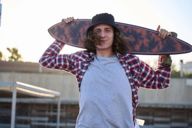 Omuzlarında bir sörf tahtası tutan genç bir kaykaycının portresi, gülümsüyor ve kameraya bakıyor.
