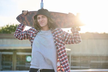 Omuzlarında uzun bir sörf tahtası taşıyan kendinden emin genç bir adam çarpıcı bir günbatımında kaykay parkının canlı atmosferinin tadını çıkarıyor.