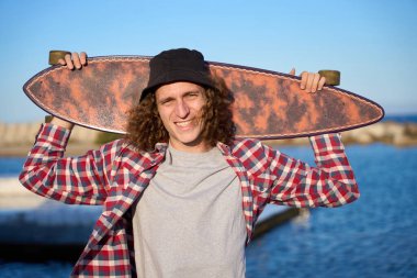 Genç kıvırcık saçlı adam kafasının arkasında uzun bir tahta tutuyor, gülümsüyor ve deniz kenarında güneşli bir günün tadını çıkarıyor.