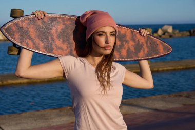 Şık bir kaykaycı, güneşli bir yaz gününde, güzel sahil manzarasında uzun kaykayını omuzlarında tutuyor.