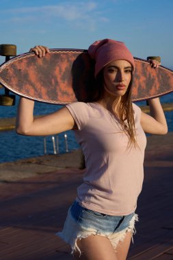 Şık bir kız rıhtımda uzun bir tahta tutuyor, parlak mavi deniz arka planda güneşli bir gökyüzünün altında uzanıyor.