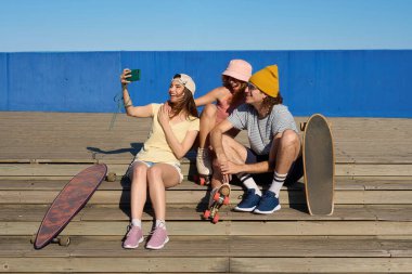 Üç neşeli genç arkadaş ahşap basamaklarda oturup akıllı telefonlarıyla kaykay ve patenleriyle selfie çekiyorlar.