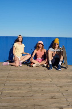 Üç havalı patenci ahşap bir iskelede, canlı mavi bir duvarın önünde güneşli havanın tadını çıkarıyorlar.