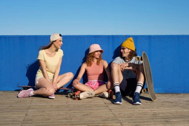 Sıradan yaz kıyafetleri giyen üç genç, tahta bir geçitte kaykay ve patenlerle oturmuş, canlı mavi bir duvara karşı konuşmanın keyfini çıkarıyorlar.