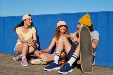 Üç mutlu arkadaş arka planda mavi bir duvarla ahşap bir geçitte oturmuş güneşli bir günün tadını çıkarıyorlar.