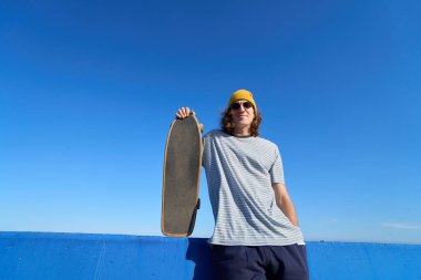 Genç patenci açık mavi gökyüzü ile mavi bir duvara karşı kaykayını tutuyor, açık havada güneşli bir günün tadını çıkarıyor.