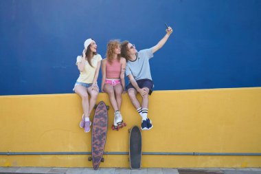 Üç genç arkadaş, kaykayları ve patenleriyle sarı bir duvarda güneşli bir günün tadını çıkarırken akıllı telefonlarıyla selfie çekiyorlar.