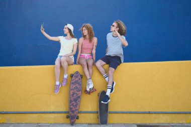 Üç genç patenci sarı bir duvarda oturmuş cep telefonlarıyla kaykay ve patenleriyle selfie çekiyorlar.