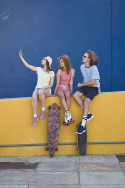 Üç havalı patenci sarı bir duvarda oturmuş akıllı telefonlarıyla kaykay ve patenleriyle selfie çekiyorlar.